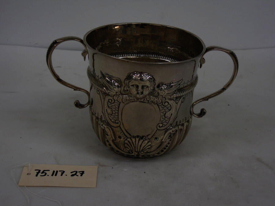 A silver cup with symmetrical handles, swirling raised designs and a small raised face on the upper half of the cup.