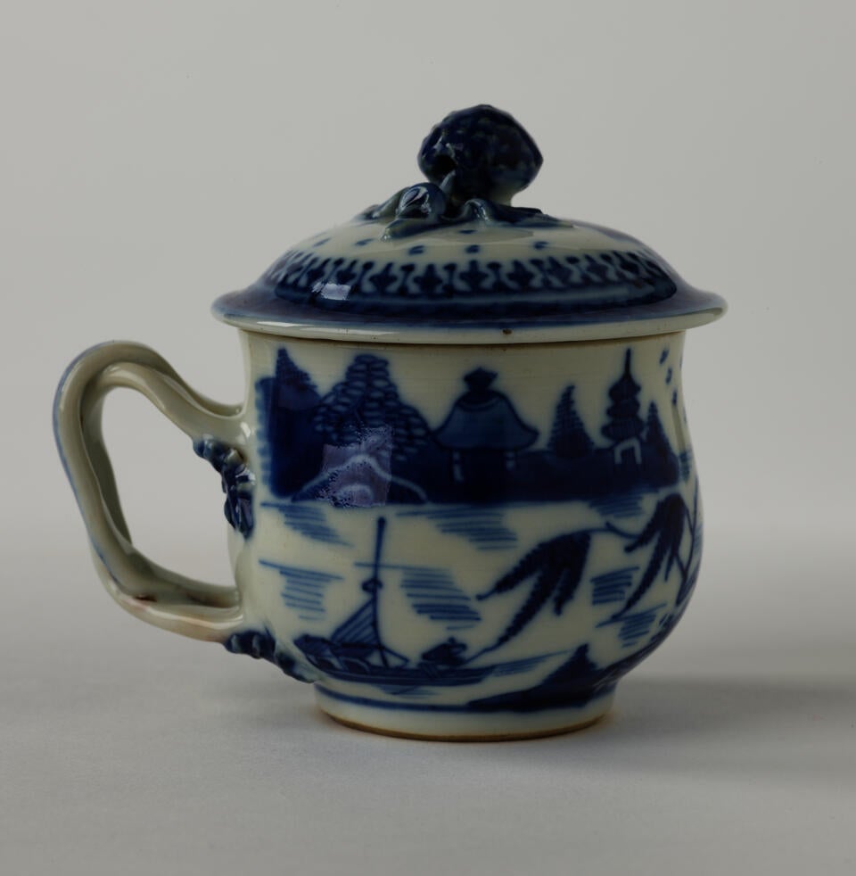 A porcelain covered cup with a sculptural swirled handle and blue architectural, floral, and scenic decorations.