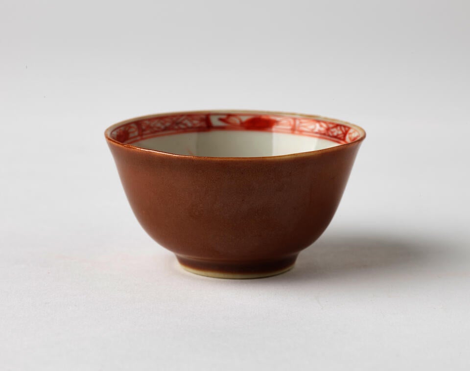 A porcelain teacup with iron-red and gold floral and abstract decorations on the interior and an orange plain exterior.