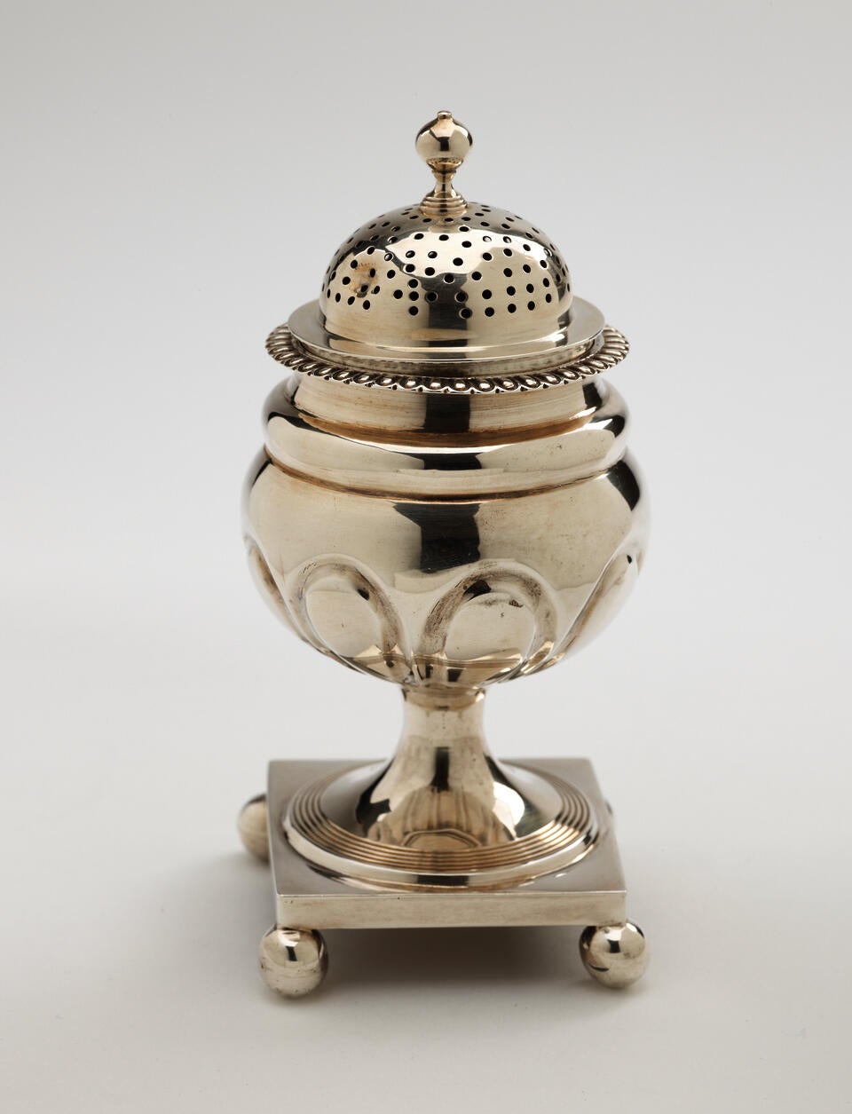 A silver caster with a square foot with small balls in each corner, the lid is perforated. 