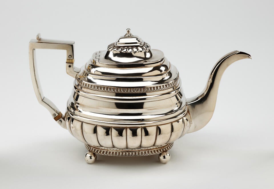 A silver luster teapot with a decorative angular handle, a rounded square body and spout with sculptural decorations.