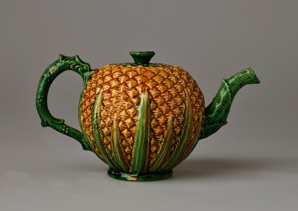 A sculptural teapot with a yellow body and texture similar to a pineapple, green handle, foot, lid, and spout with a floral leaf or stem like decoration and texture.