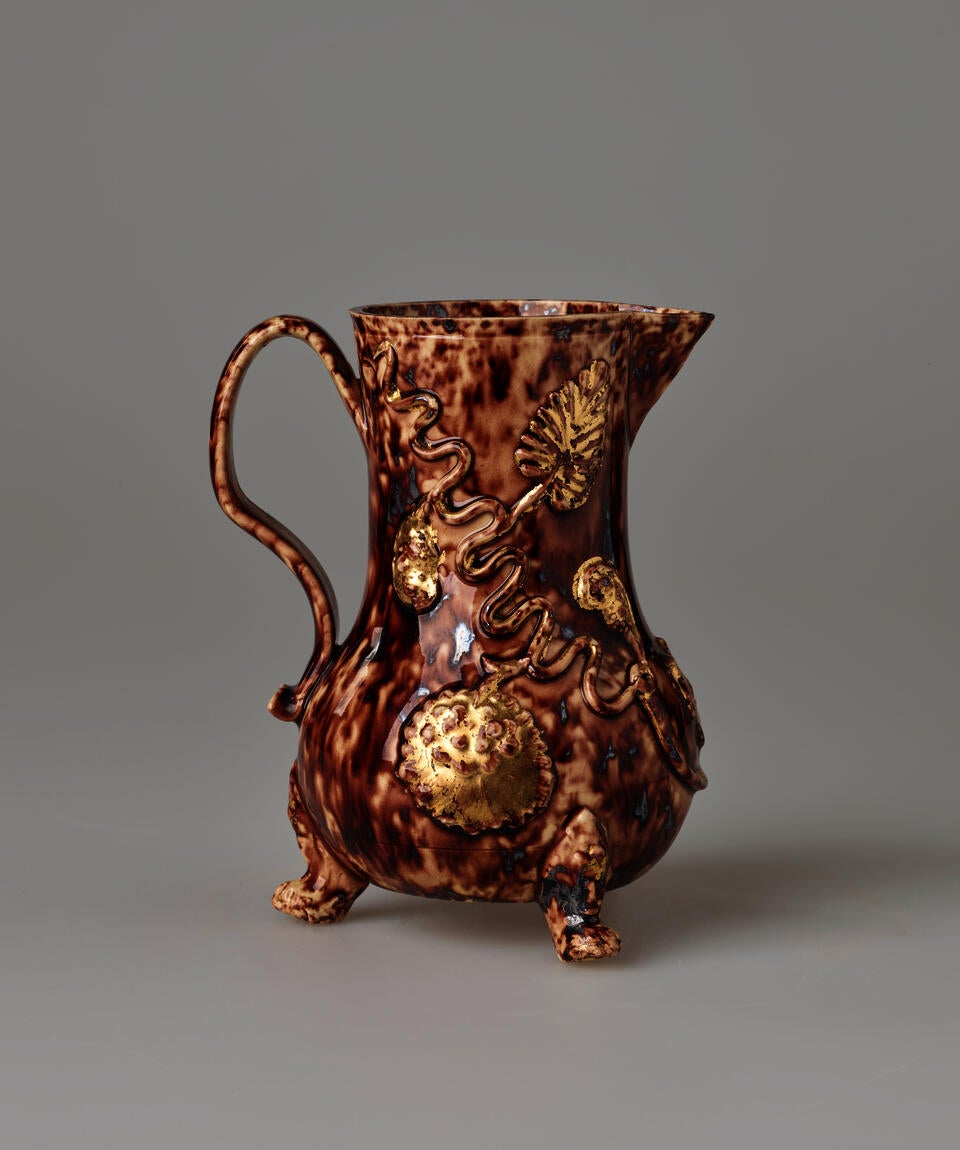 A brown ceramic pitcher with a spout, handle, and multiple delicate feet. There are swirly floral decorations.