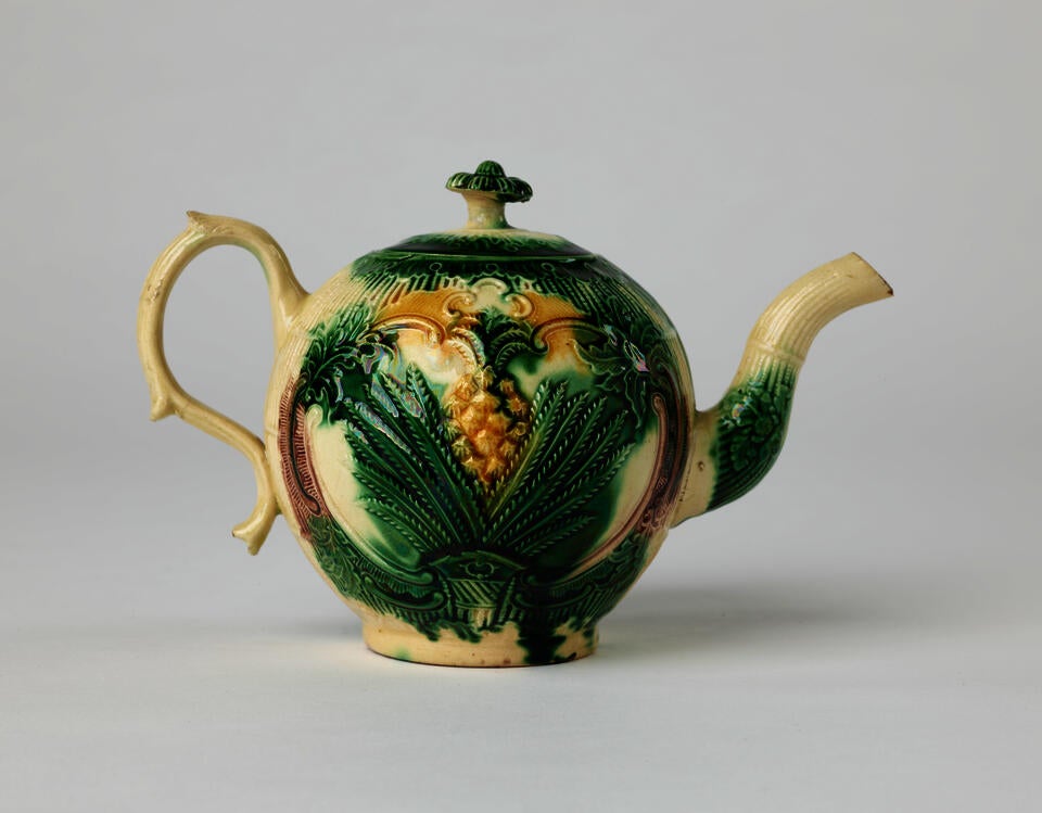 A bulbous ceramic teapot with a long curved spout and handle, white, dark green. and yellow glaze, and floral decorations ranging from leaves to a more central pineapple.