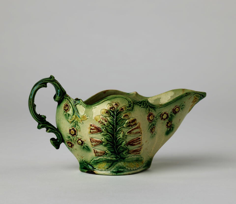 A short boat shaped pitcher green, cream, and brown in color with a sculptural handle and floral decorations.