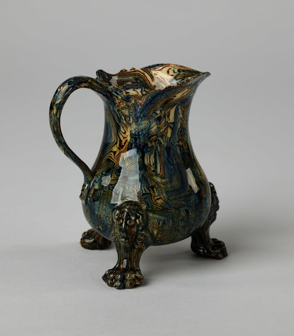 An agate wear vessel with a handle, three sculptural feet, and a spout. The vessel is shiny and has cream, blue, and green swirling colors.