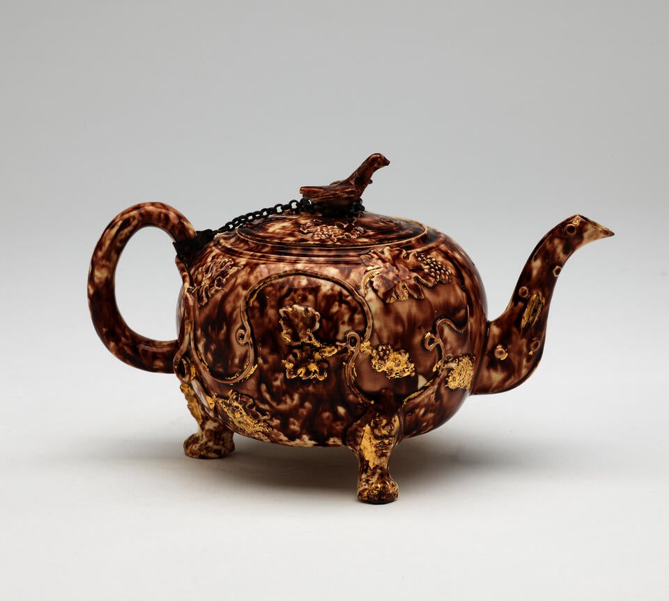 A dark brown and white ceramic teapot with protruding feet, a spout, handle, and lid. The finial on the lid is connected to the handle by a chain.