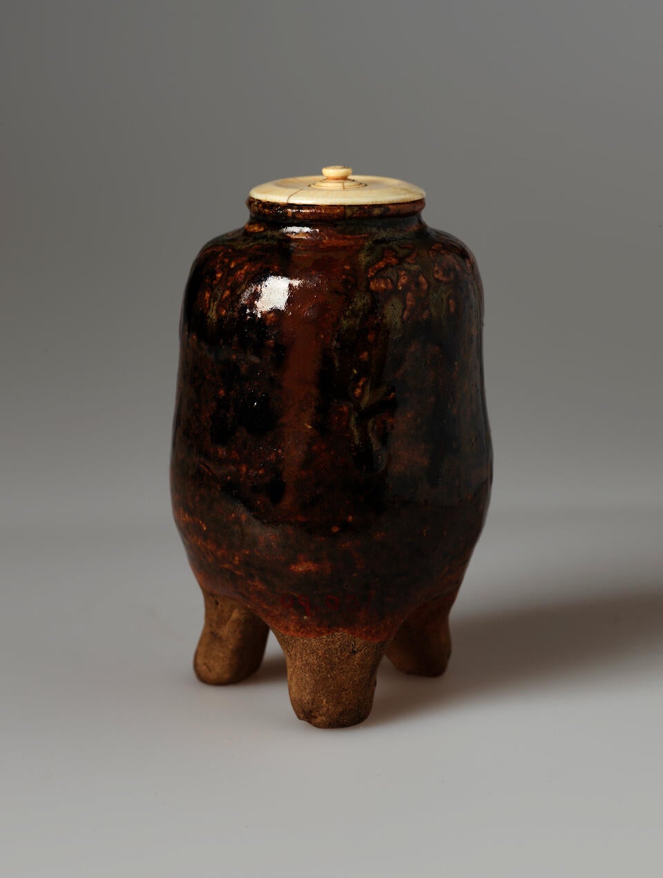 Small vessel in shades of reddish brown and black, standing on three rounded feet, with a cream colored lid.