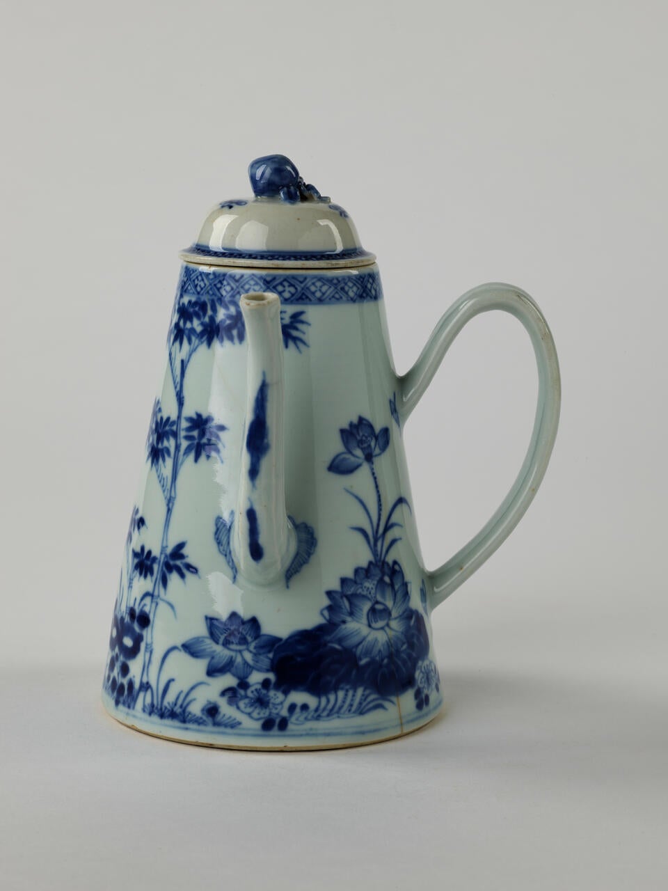 A white and blue teapot with floral decorations, a lid with a small semi spherical finial, spout, and a handle which is located approximately 90 degrees from the spout.