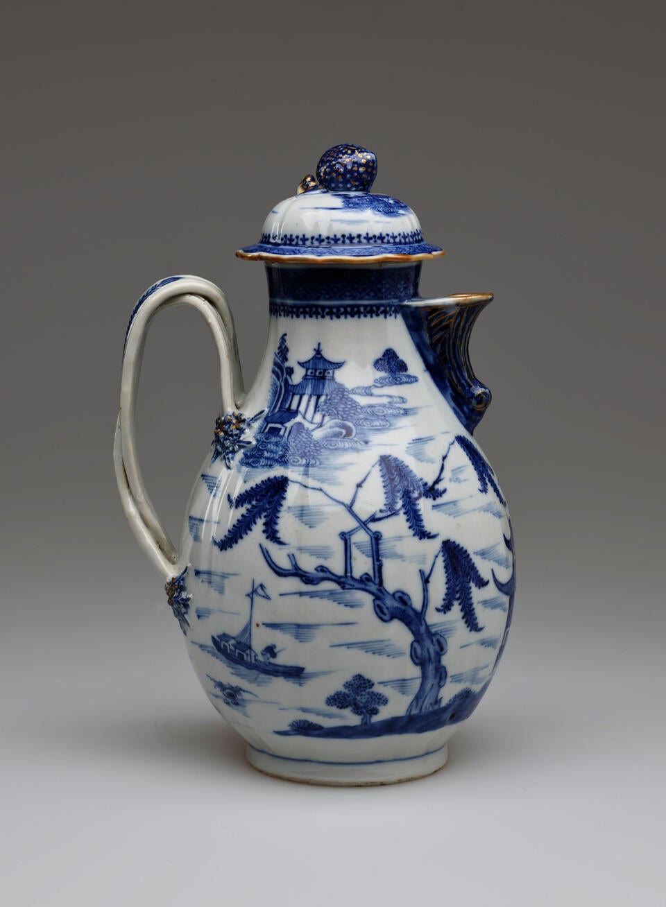 A white and blue chocolate pot with a spout, handle, and lid, the edges of the lid and spout are gilded. The decorations mainly depict floral, architectural, and landscape elements.