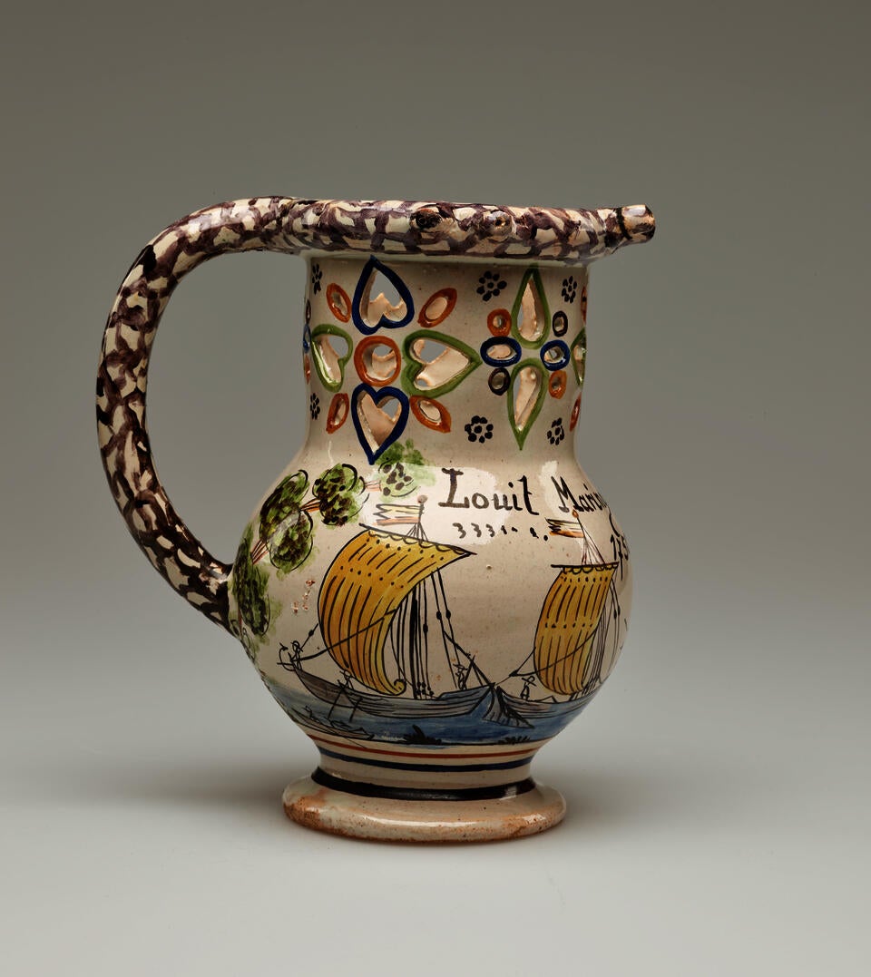 A jug with a rounded handle and pierced designs. The cream colored body is decorated with images of ships and a tree. 