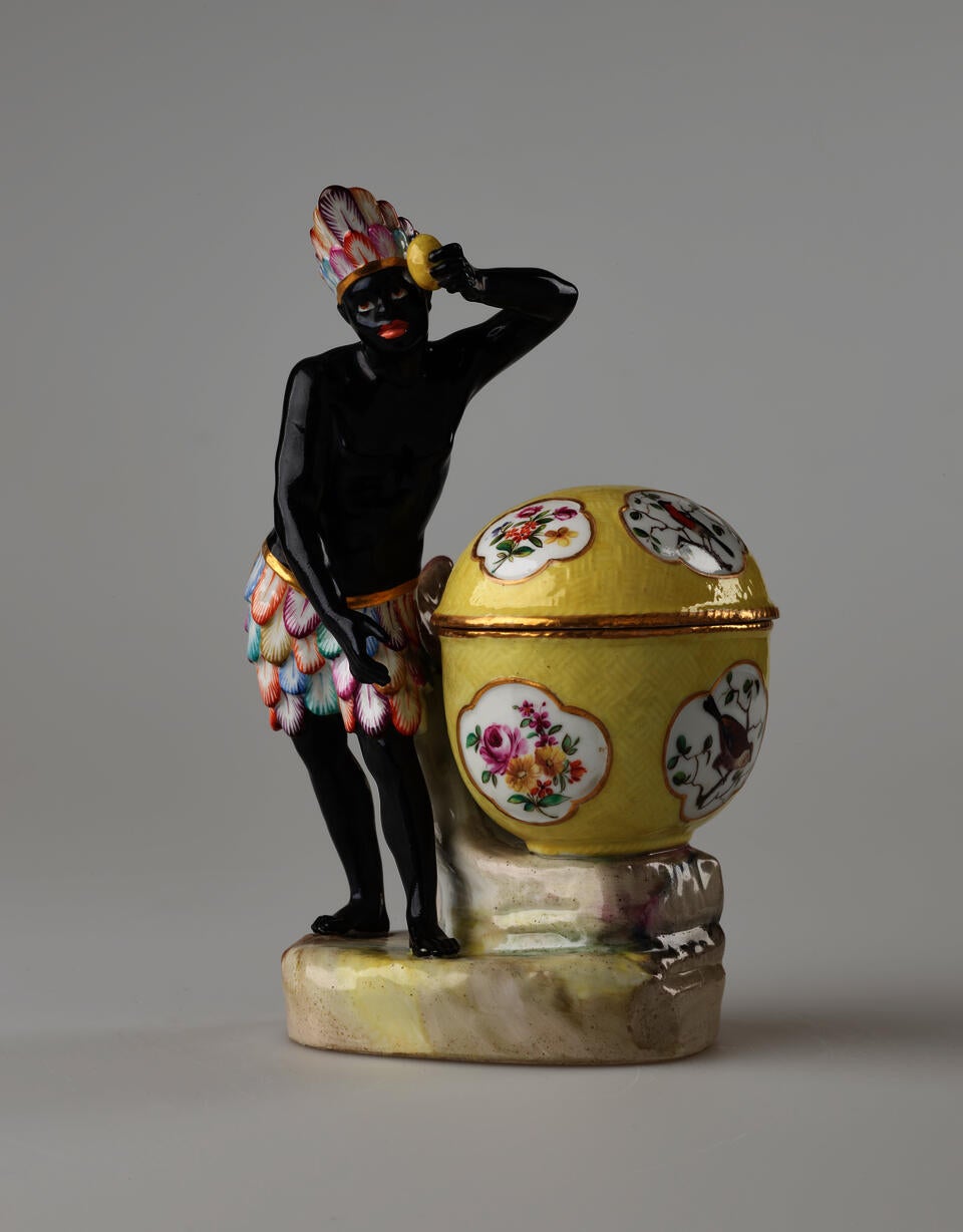 A sculptural figure with glazed black skin, a colorful headdress and bottoms. This figure is standing next to a bright yellow covered sugar basket with floral decorations. 