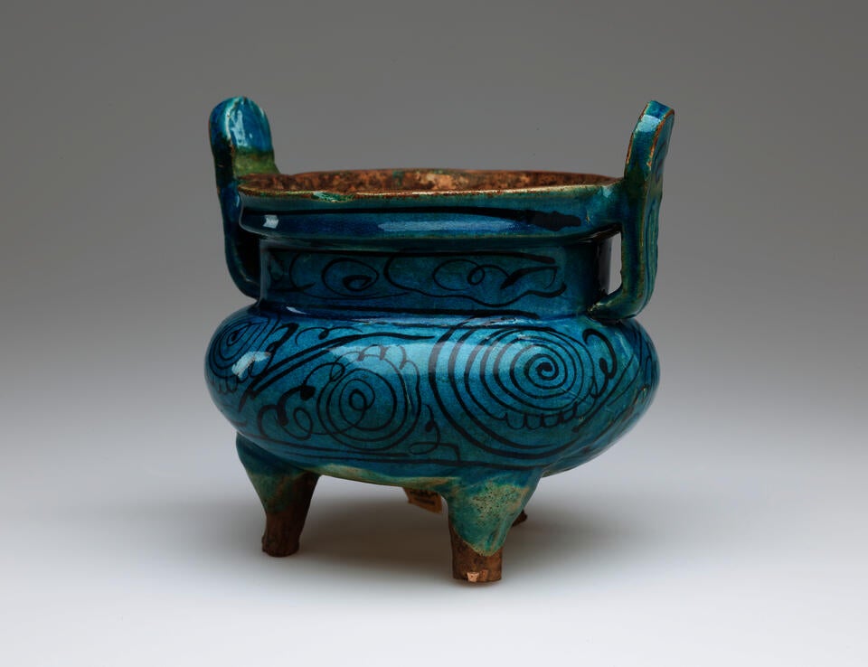 BP- Round, blue vessel with dark thin detailing on body. Two thin handles flare upward, and has four unglazed feet. 