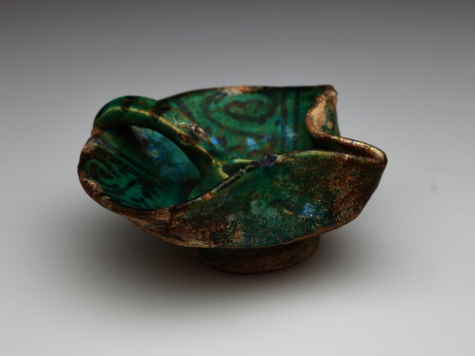 An irregularly shaped blue-green iridized vessel with dark designs. Rounded on one side with a handle that comes to the center of the vessel. The other side has a folded spout-like edge.