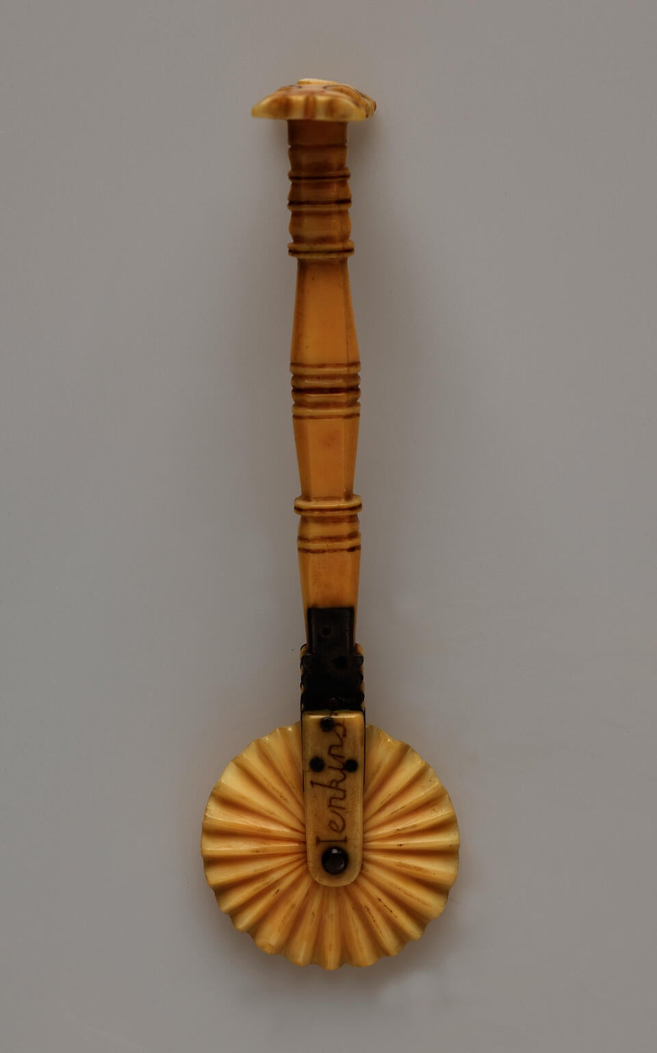 An ivory pie crimper with a decorative handle and a textured wheel. There are letters on the part attaching the wheel to the handle.