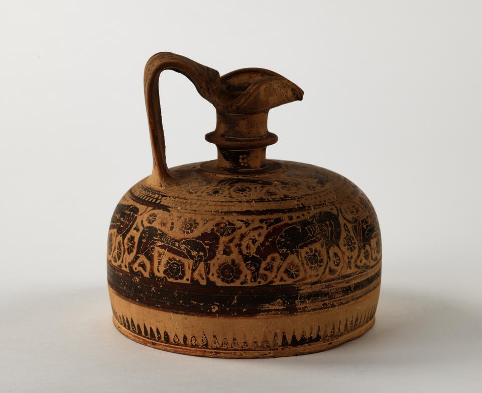 A terracotta wine jug with a thick handle, wide cylindrical base, smaller neck, a folded rim and brown and black decorations that are worn and depict abstract shapes and animals. 