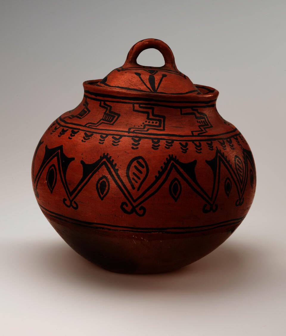 A bulbous red vessel with black abstract designs, leaning slightly to the right. The lid has a small round handle. 