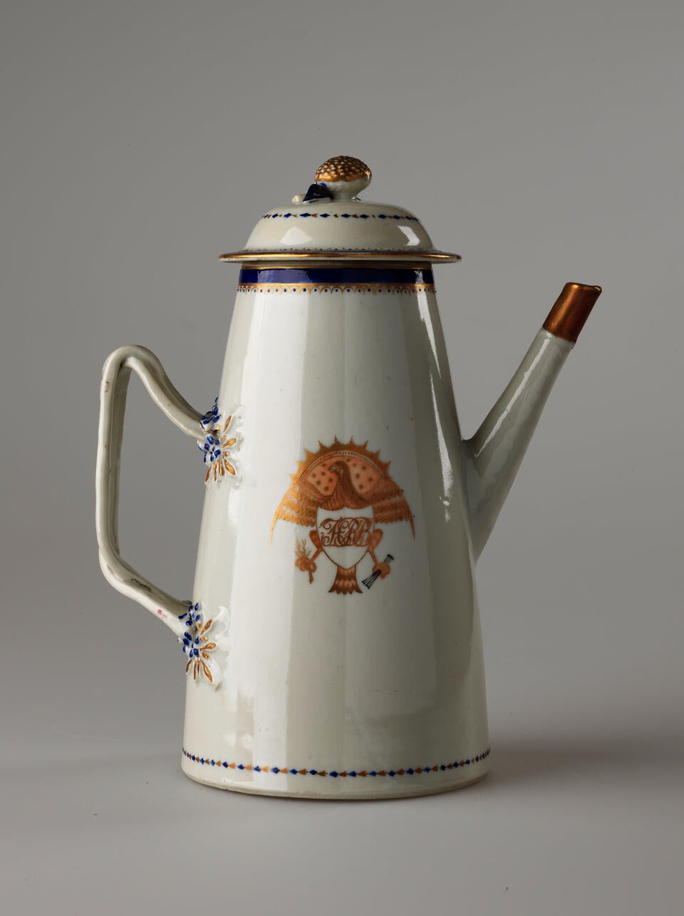 A white, blue and gilded teapot with a long straight spout, a handle, and lid. The central decoration depicts a heraldry bird with a shield and objects in its talons.