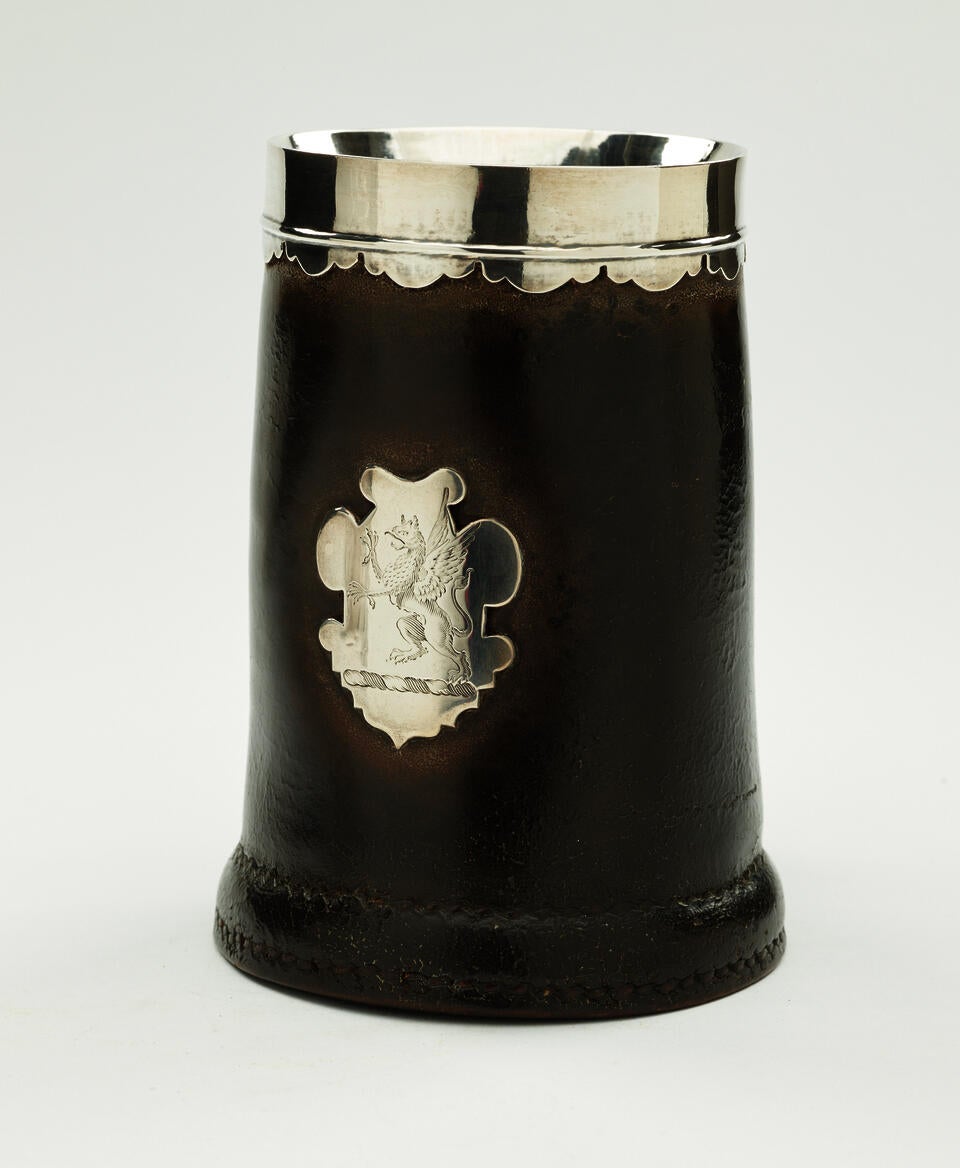 BP: A leather, silver, and pewter tankard. The body is mostly dark with the exception of a decorative crest, rim and interior. 