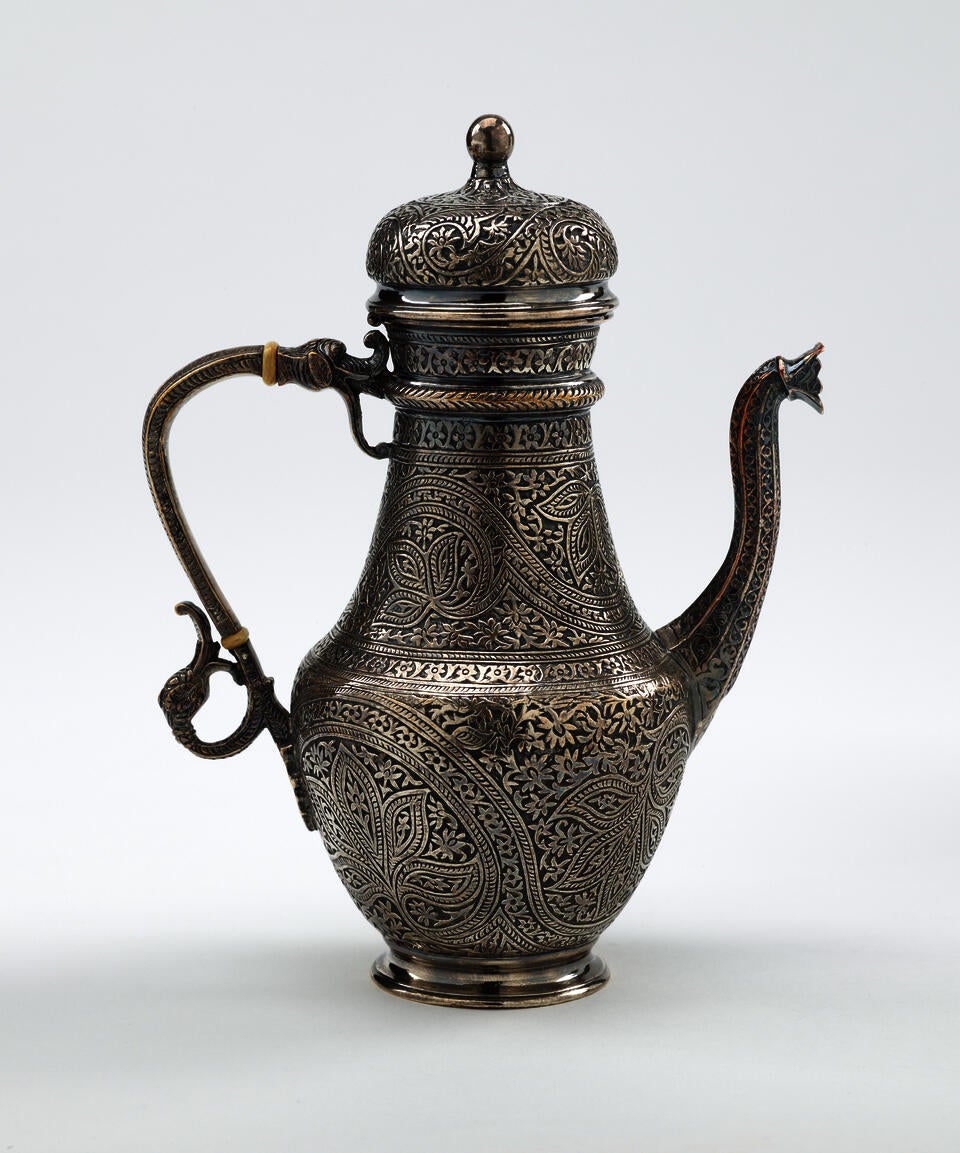 A silver and ivory coffee pot with detailed delicate decorations, handle with a lid hinged to it, and spout.
