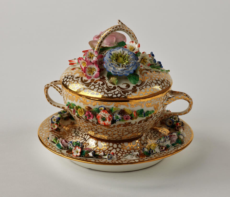 A porcelain two handled covered bowl and fitted saucer. The decorations are gold, pink, green and are floral. 