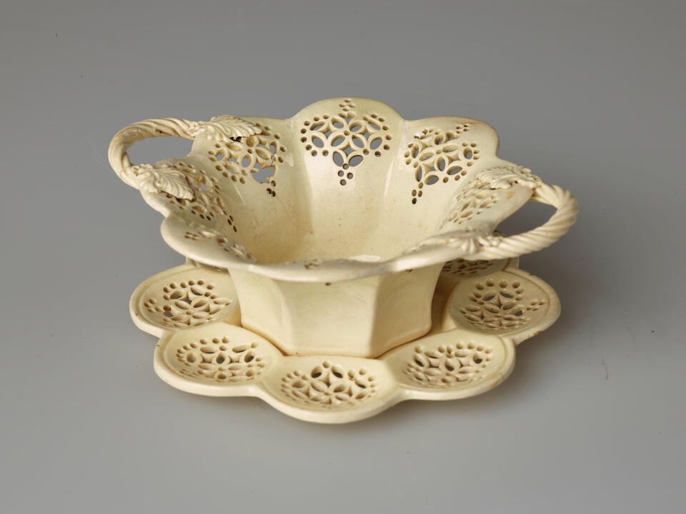 A creamware sweetmeat dish with swirled handles and lace-like decorations cut into the octagonal vessel that is sitting on a matching tray.