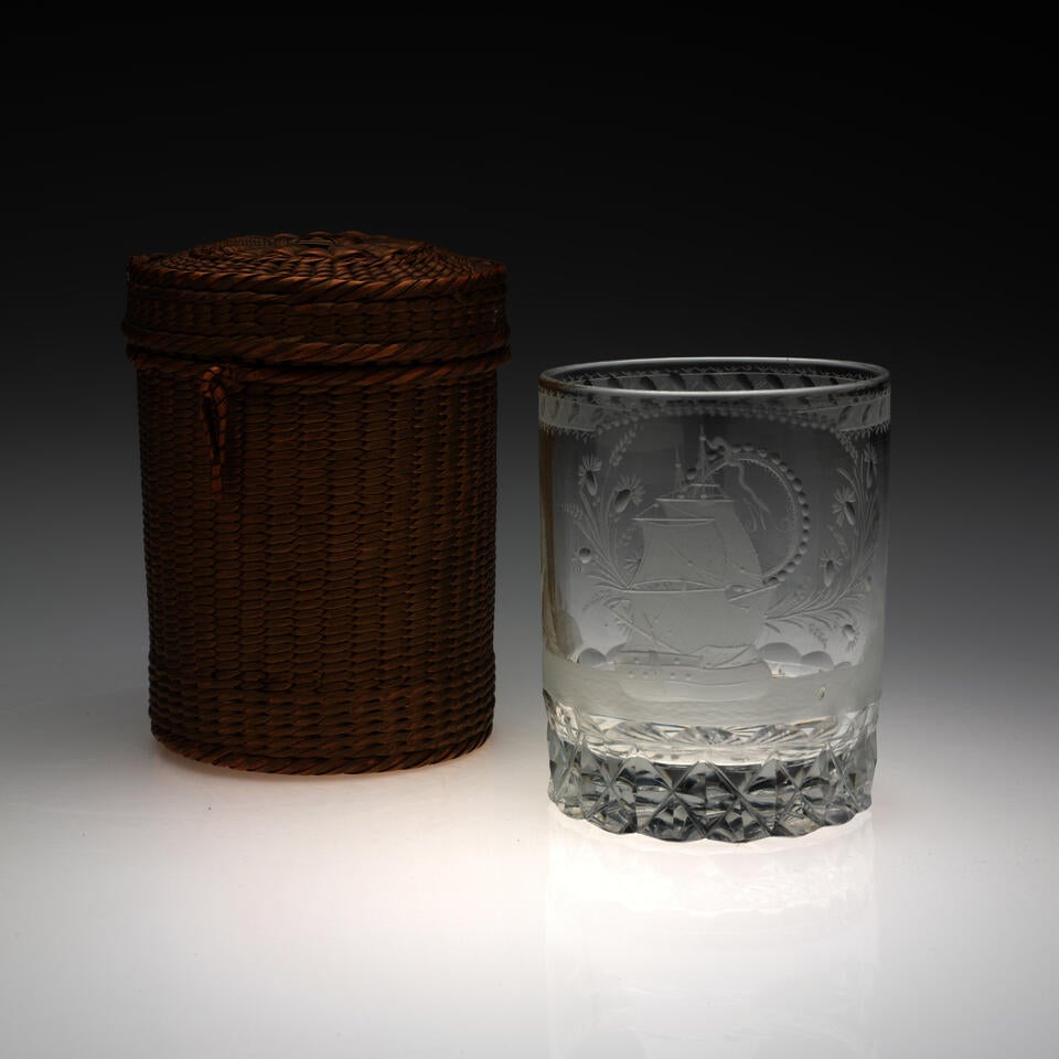 Rounded clear glass with etched designs next to a brown woven basket of the same shape. A small, tan-colored implement lays to the left of the basket and glass. 