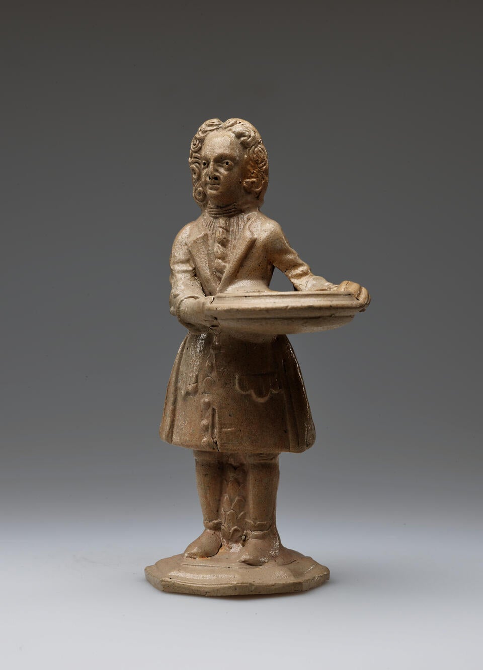 A sculptural figure standing, holding a vessel, dressed in historical clothing with tightly curled hair. The entire stoneware figure is a light cream color.