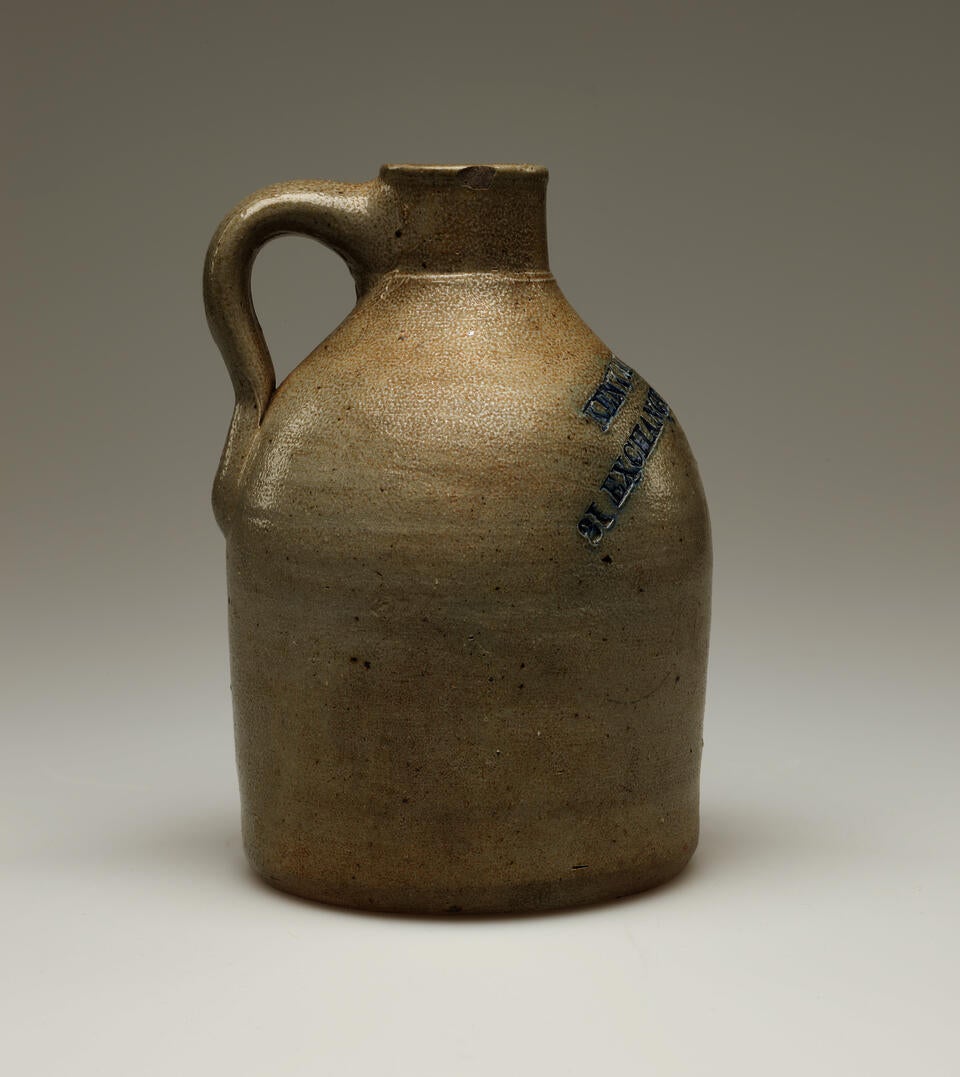 A round, sand colored jug with a small, rounded handle. Blue colored text  is stamped into the body of the jug, on the left side. Legible text reads, “PROV RI.”