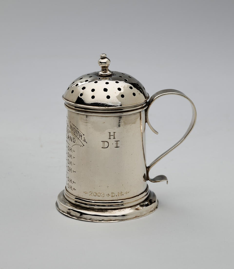 A container with a rounded handle, pierced lid, and small rounded finial. The letters “HDI” are engraved on the right side and a list of text is engraved on the left side. 