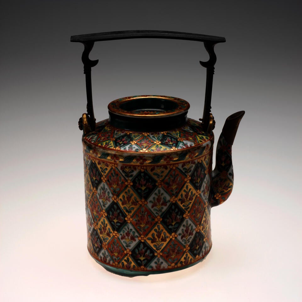 A cylindrical teapot with an elaborate design of flowers inside diamond shapes of varying colors. A thin, square shaped handle with a flat top sits above the teapot.
