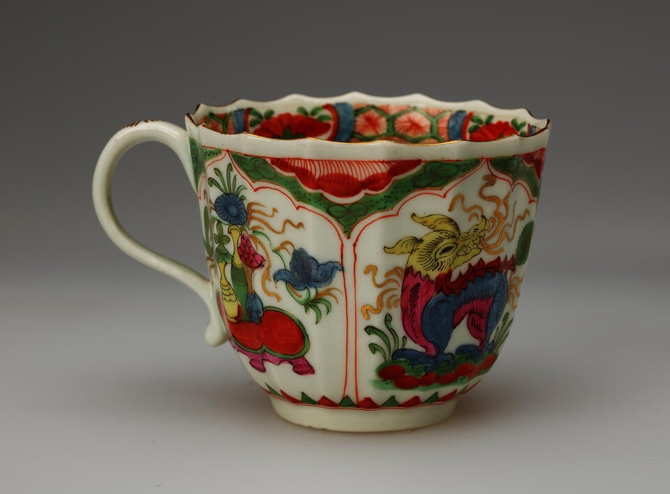 A white cup with a scalloped lip, green, red, blue, pink, yellow, gilded decorations, and two vignettes: an animal and the other a table with vessels on top of it.