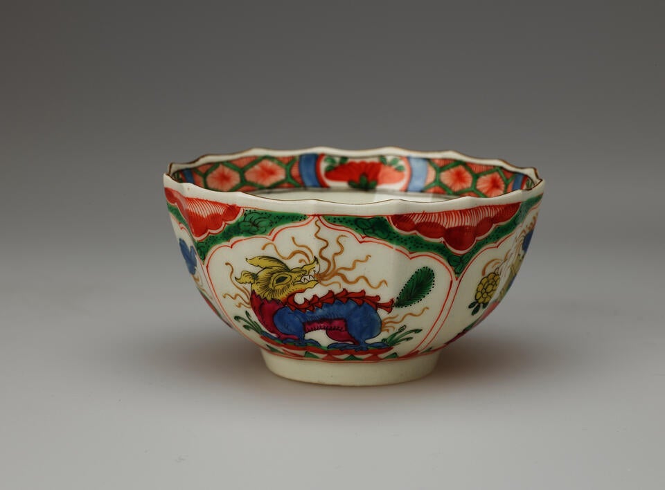 A white tea bowl with green, red, blue, pink, yellow and gilded decorations. The lip of the vessel is scalloped. 