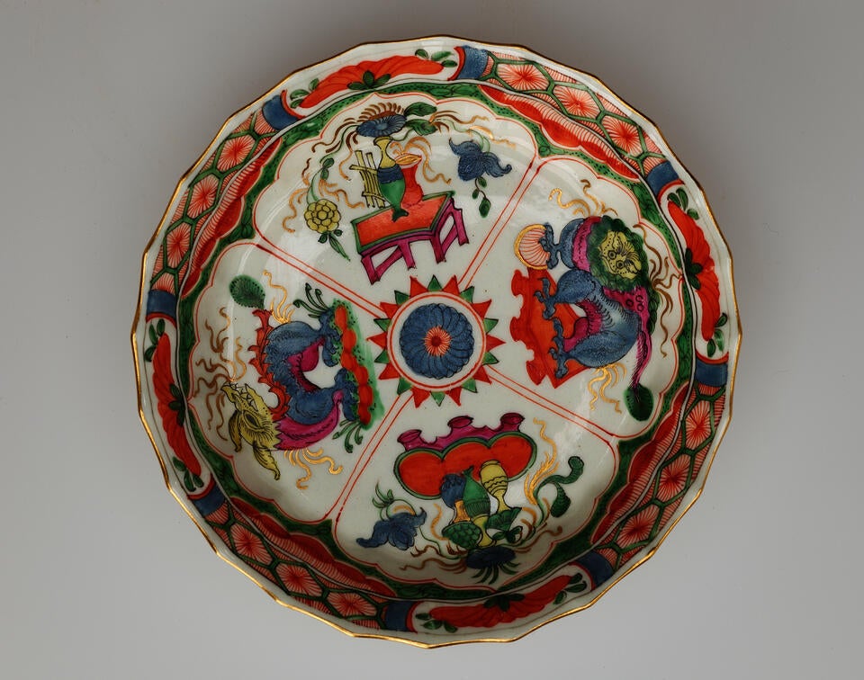 A white saucer with green, red, blue, pink, yellow, and gilded decorations; separated into four main vignites, two animals and two tables and vessels containing floral elements.