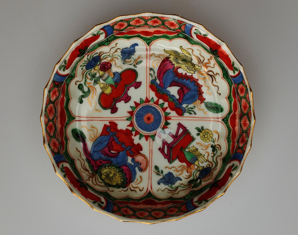 A white saucer with green, red, blue, pink, yellow, and gilded decorations. Decorations separated into four vignettes; two animals and two scenes of a table and vessels containing floral elements.