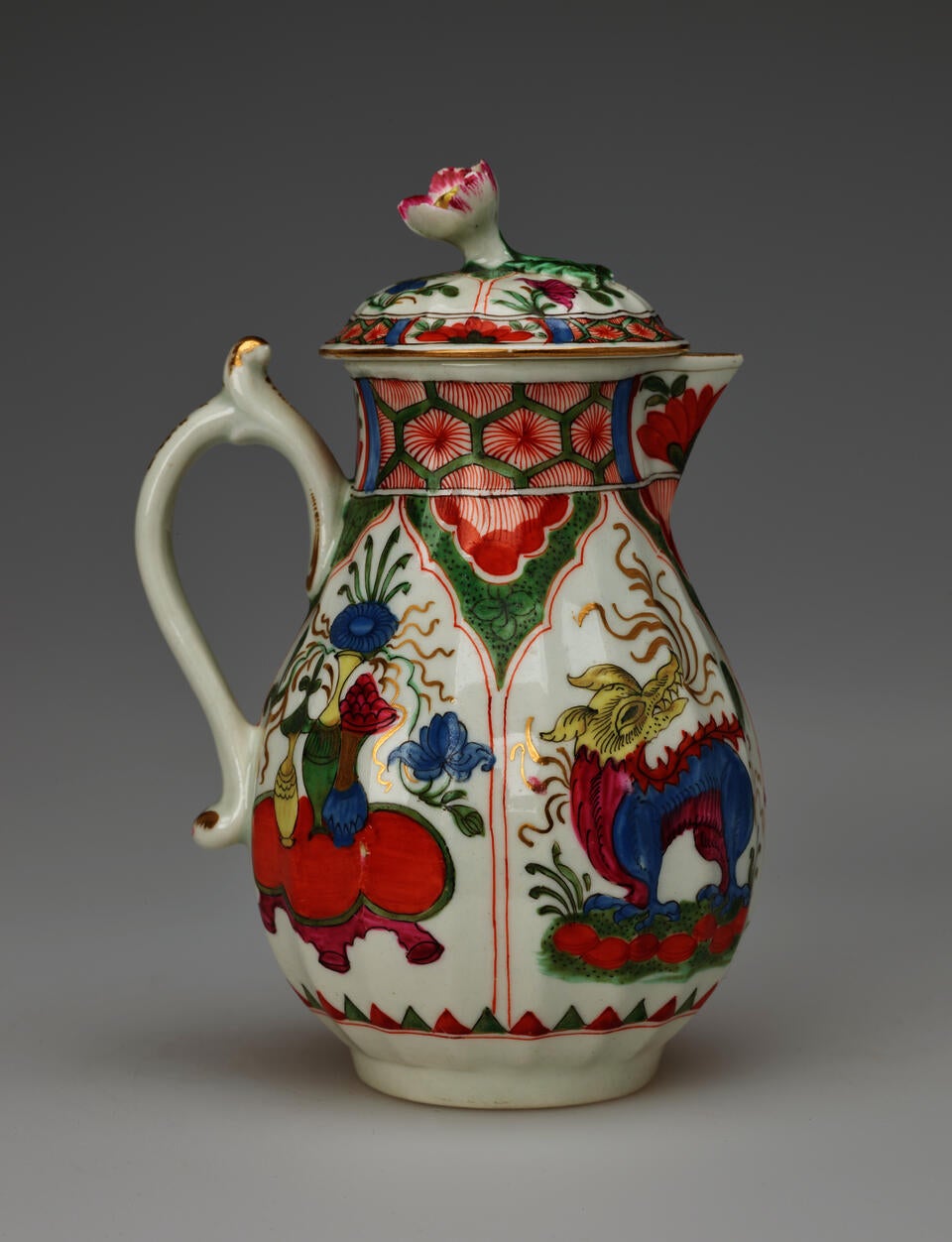 A white milk jug with a handle, lid, and spout with green, red, blue, pink, yellow, gilded decorations,and two vignettes; an animal and a table, vessels, and floral elements.