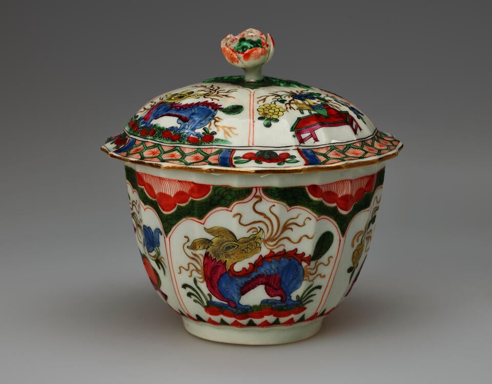 A white sugar bowl with lid and finial with green, red, blue, pink, yellow and gilded decorations. The finial is a sculptural flower white, red and green.