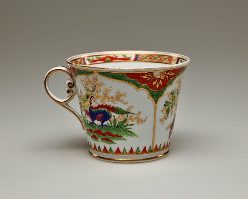 A chocolate cup that is with green, red, blue, pink, yellow, gilded decorations, and vignette with a dragon on a table surrounded by gilding.