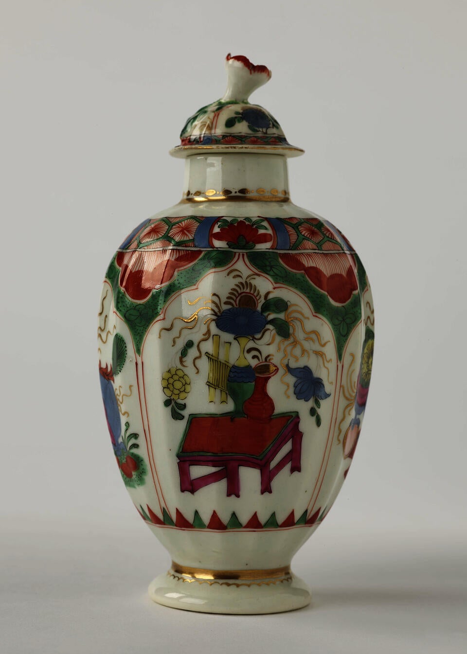 A porcelain tea caddy with a lid and green, red, blue, pink, yellow and gilded decorations. The finial is a sculptural flower white, red and green