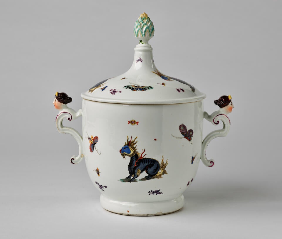 A white porcelain icecream pail with dragons and bugs decorations, there are two sculptural handles that are figure’s heads, the lid has a sculptural finial.