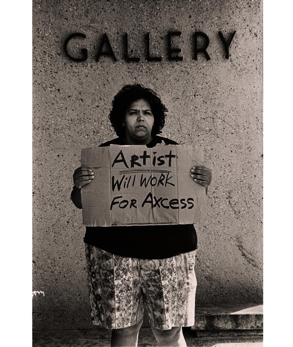 Black and white photo of an unsmiling woman looking at the camera and holding a sign that says “Artist. Will work for axcess.” Lettering on the wall above says “Gallery.”