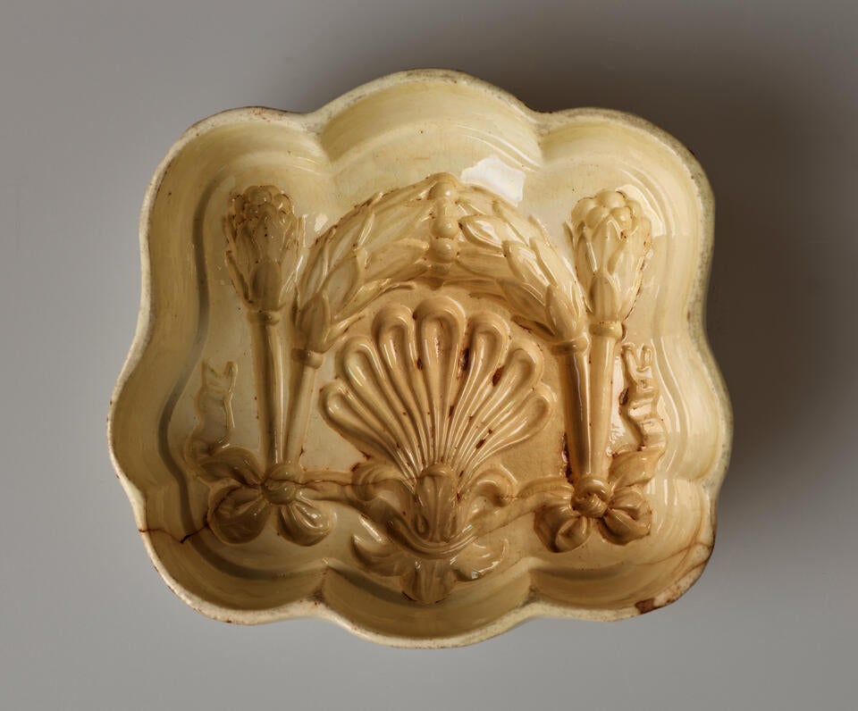 A ceramic pudding mould that is cream colored and is curved with inset floral and bow decorations.