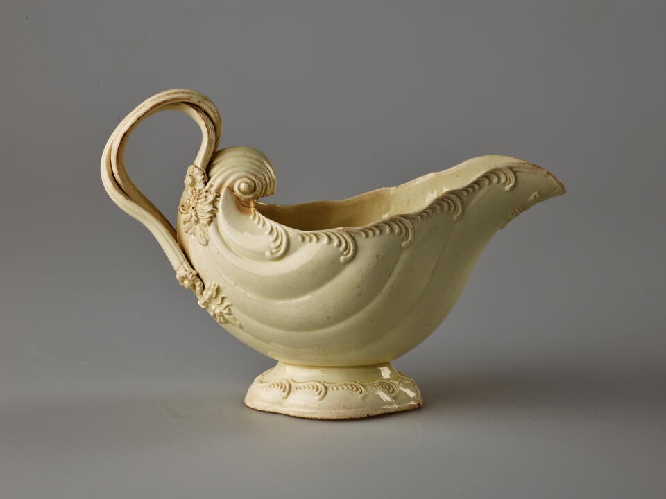 A cream colored vessel with rounded foot and handle. The body of the vessel is shell shaped and has swirled decorations. 
