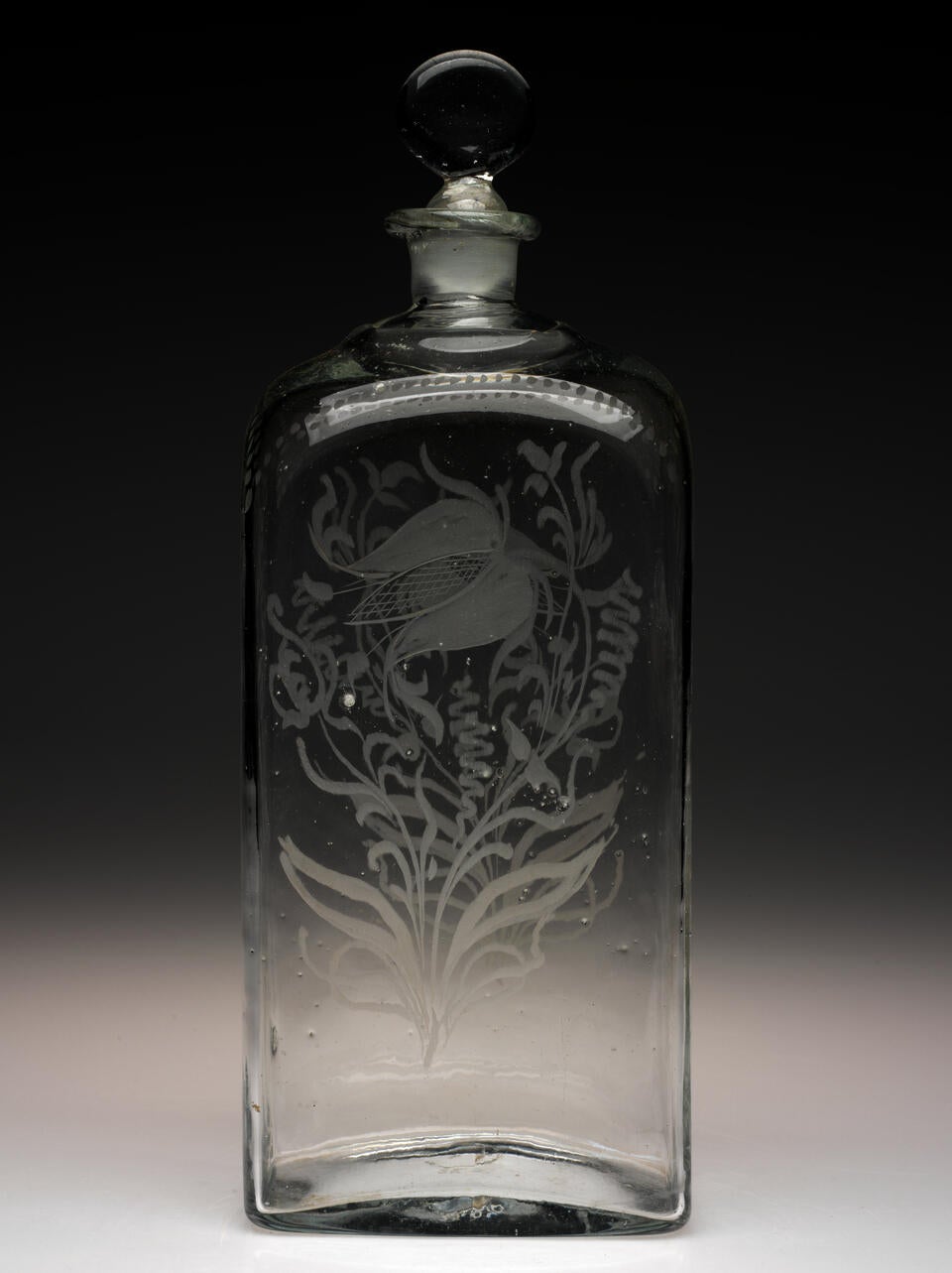 Tall rectangular clear bottle with rounded stopper. A floral design with leaves and swirling lines is etched on the front and back. 