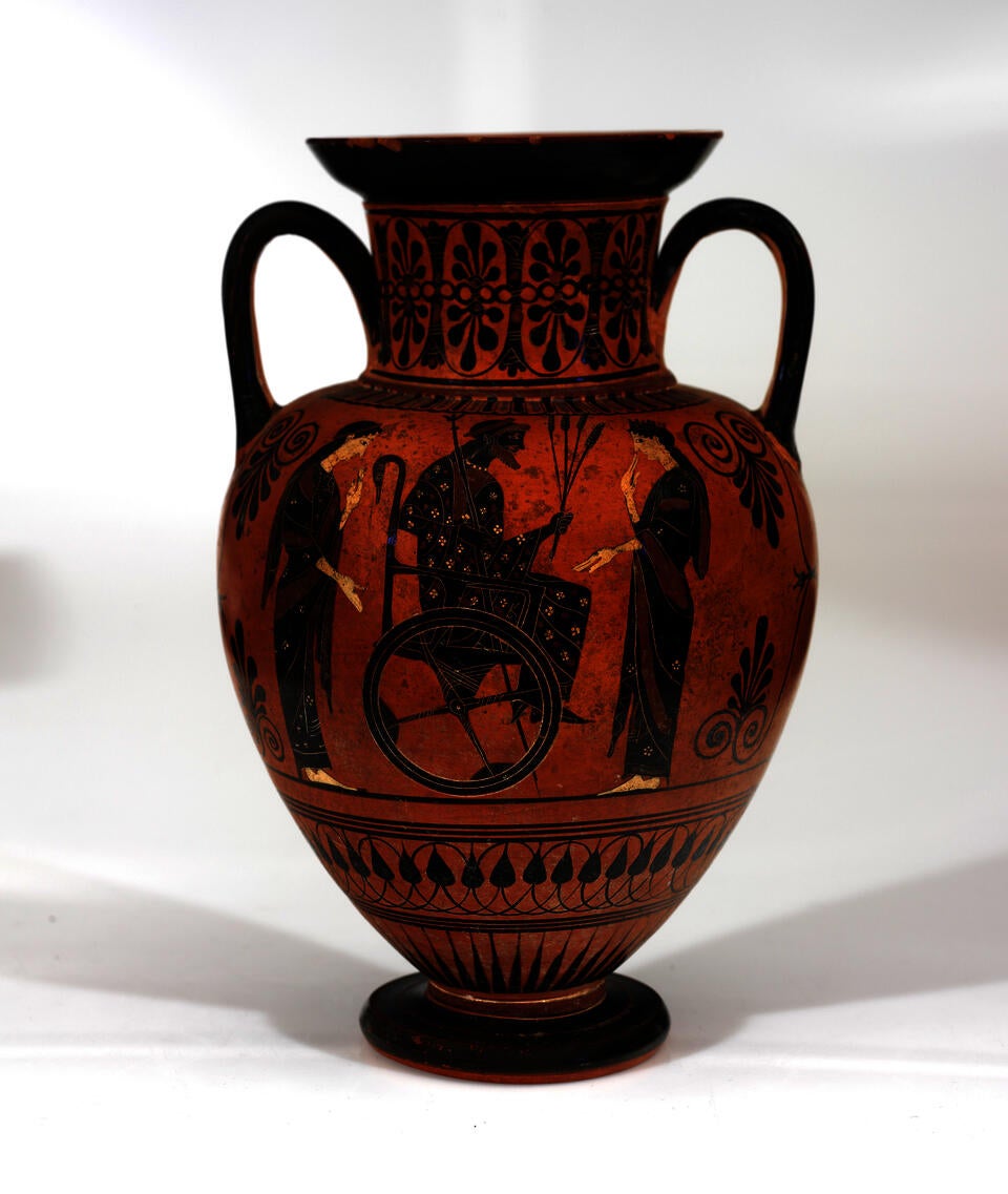 Orange clay jar with two handles. It is decorated with black patterns and an illustration of a man in a wheelchair holding a staff and grain, flanked by two women.