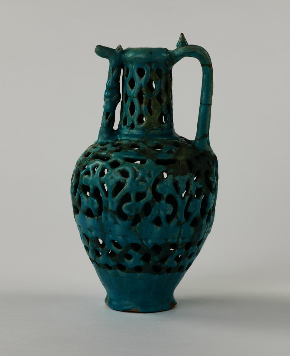 A turquoise earthenware ewer with cut out decorations and a sculptural spout and handle.