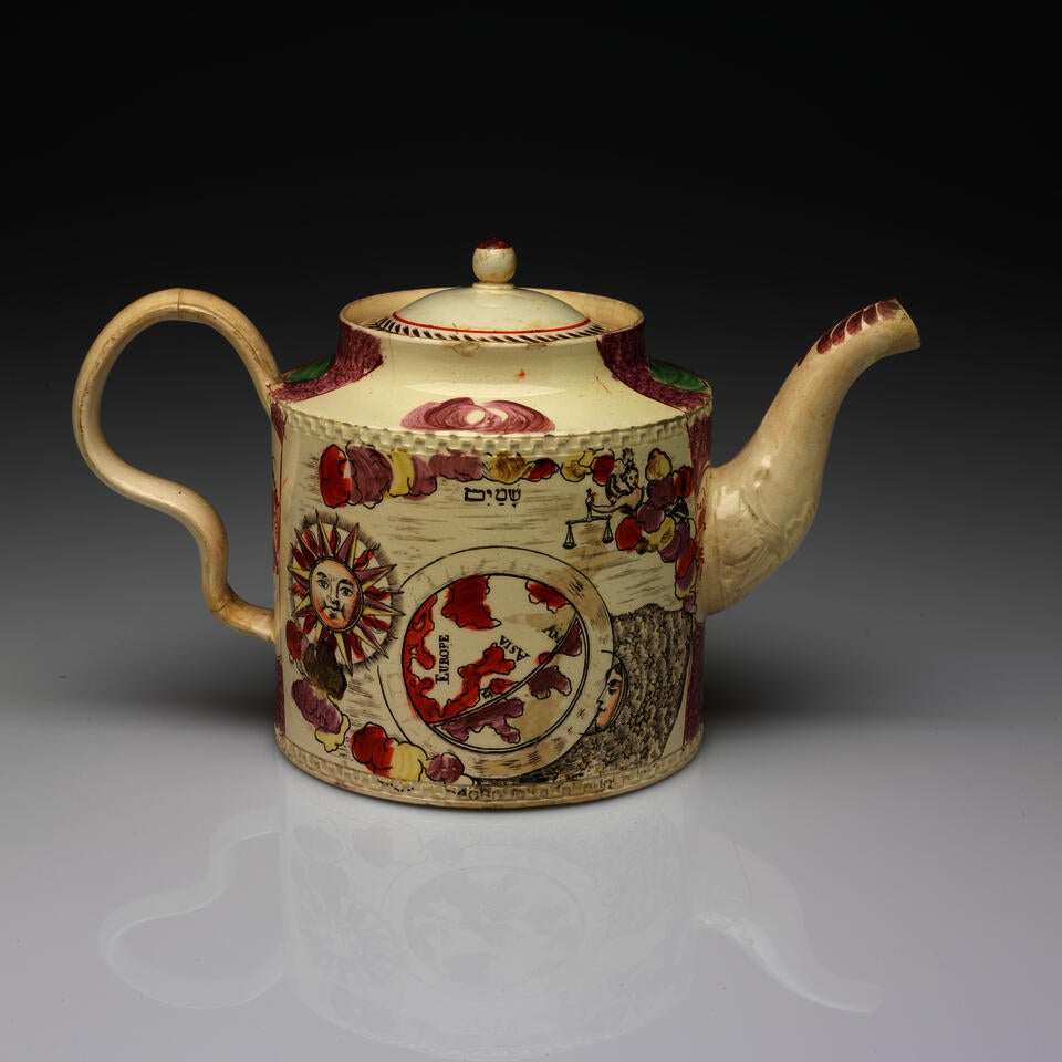 A cylindrical teapot with an inward curve towards the lid, a wavy handle, and a spout. The transfer printed decorations are a sun, a map, and a figure holding scales.