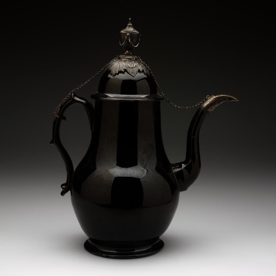 A glazed black teapot with a decorative handle, long curved spout, and a short foot, metal decorations on the spout, lid, finial, and handle with a chain connecting them. 