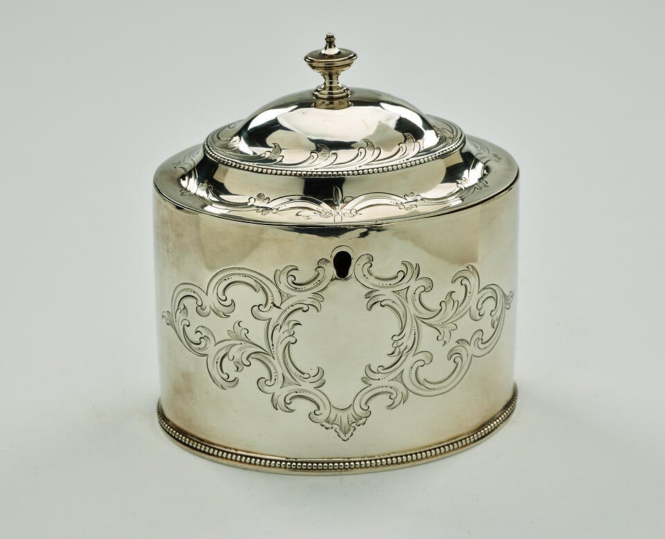 A silver tea caddy with a lid and sculptural finial. There is also swirling engraving along the body and lid.  