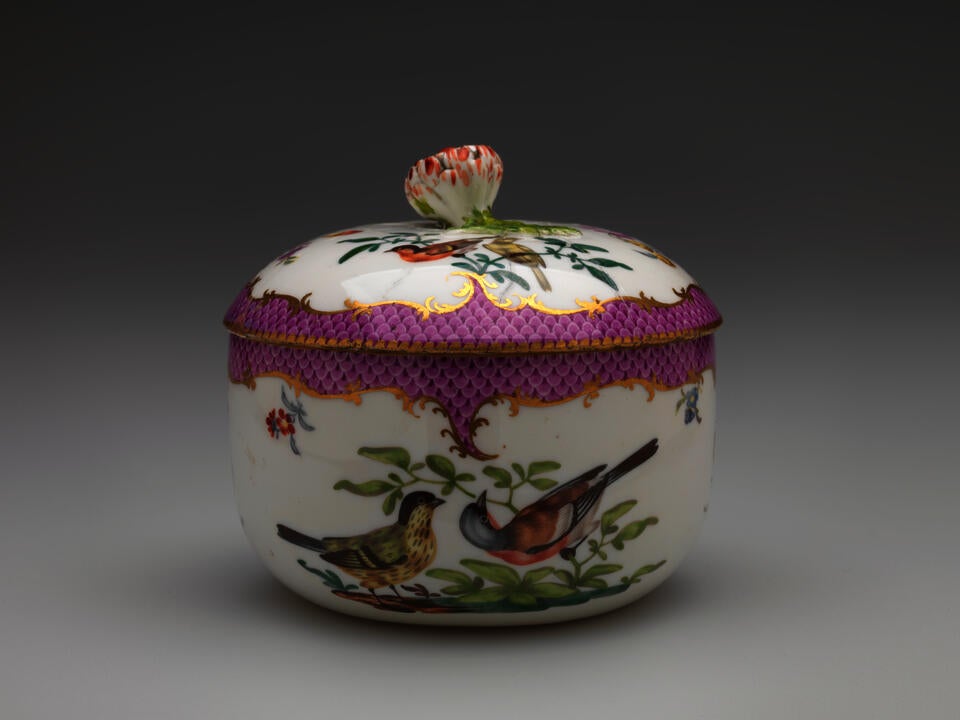 A white vessel with a lid, with  gilded and pink decoration, floral decorations, and two birds that are white, black, red and blue, both are on a branch.